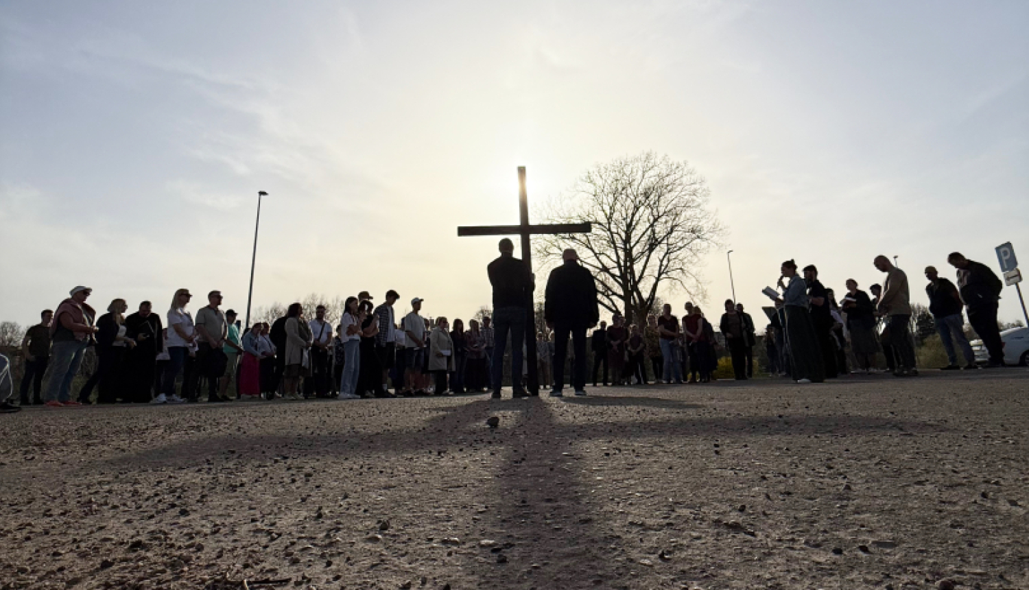 Zema rakursa fotogrāfijā redzams cilvēku loks brīvā dabā ap lielu koka krustu. Priekšplānā redzams grants segums, bet fonā – cilvēku silueti pret saules gaismu, kas atrodas aiz krusta. Pie krusta stāv divi cilvēki, apkārt pulcējušies vairāki desmiti cilvēku. Fonā redzams arī koks un skaidras debesis.