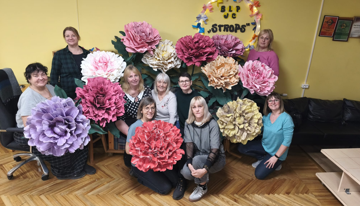 Grupas foto redzamas desmit sievietes Brīvā laika pavadīšanas un jauniešu centra „Strops” telpās, pozējot ar lielformāta papīra ziediem dažādos toņos – rozā, baltā, ceriņkrāsas, sarkanā, krēmkrāsas un dzeltenīgā. Fonā pie sienas redzama centra dekorācija ar uzrakstu „BLP JC “Strops””, bet telpā ir dzeltenas sienas, koka grīda un tumšs dīvāns.