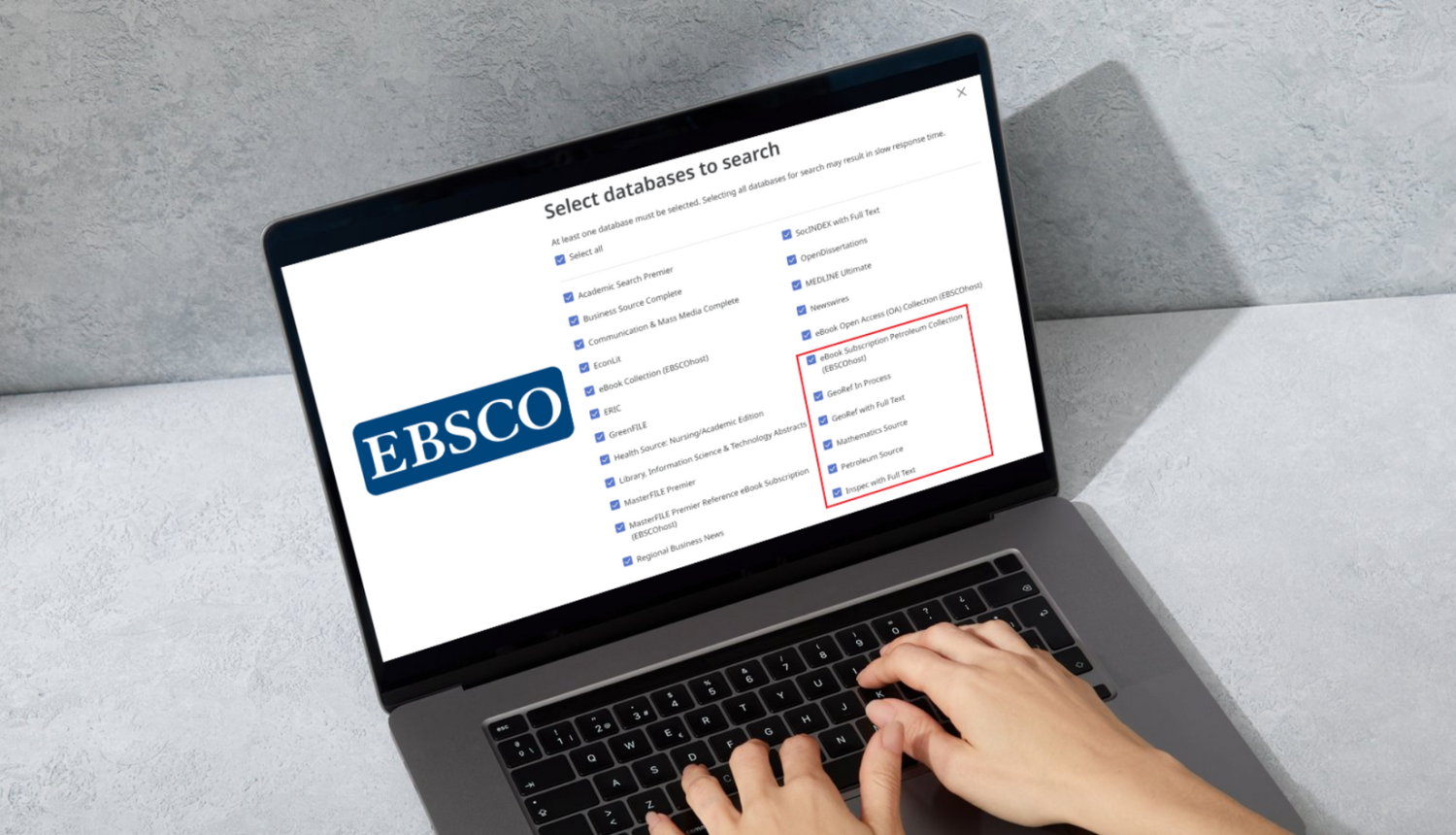 Klēpjdators ar atvērtu EBSCO datubāzu atlases ekrānu („Select databases to search”); redzams EBSCO logotips un ar sarkanu rāmi izcelta datubāzu sadaļa, priekšplānā – cilvēka rokas uz tastatūras.