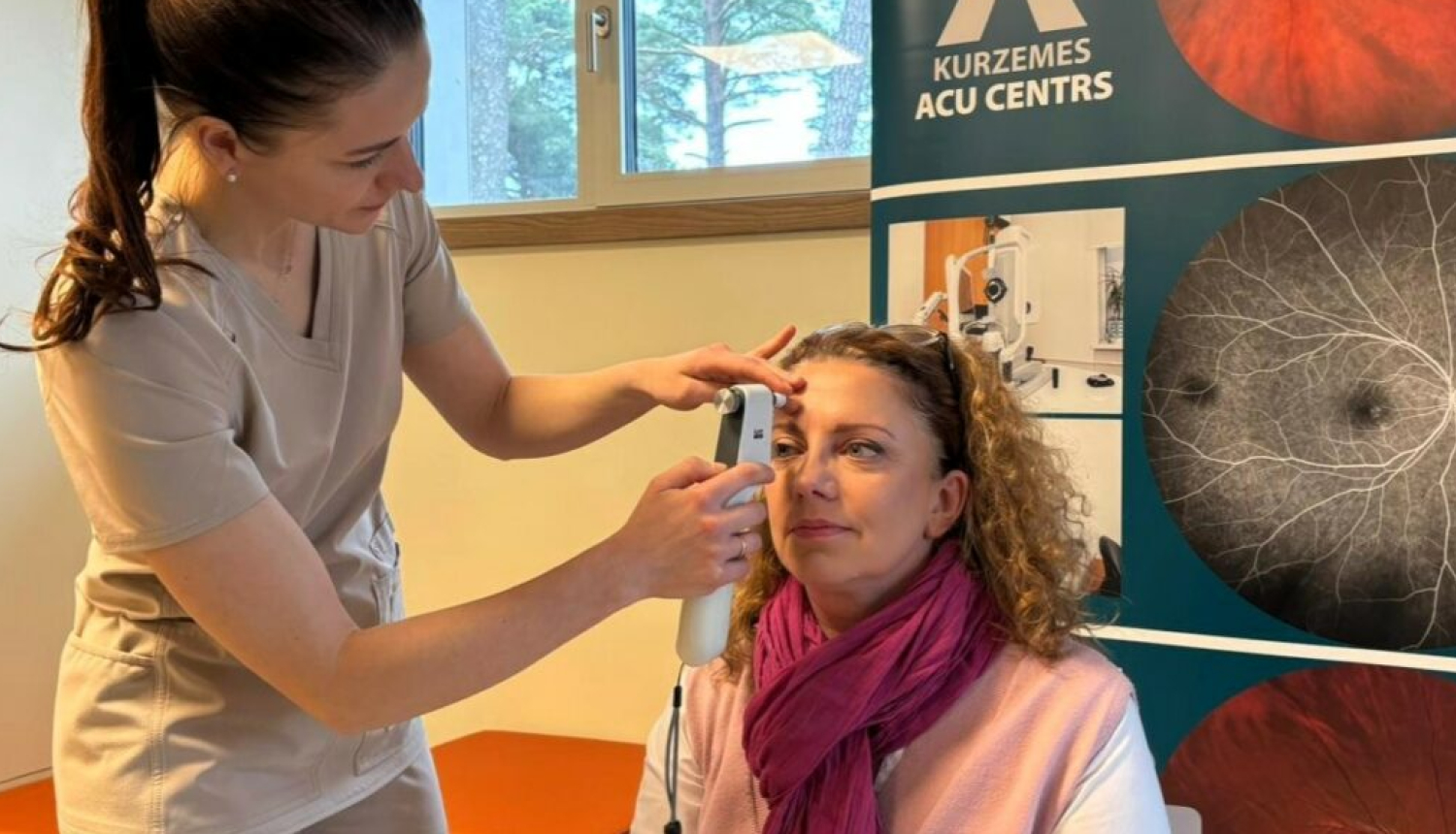 Medicīnas darbiniece veic acu spiediena mērījumu sievietei, kura sēž krēslā, bet fonā redzams “Kurzemes acu centra” stends ar acu izmeklējumu attēliem.