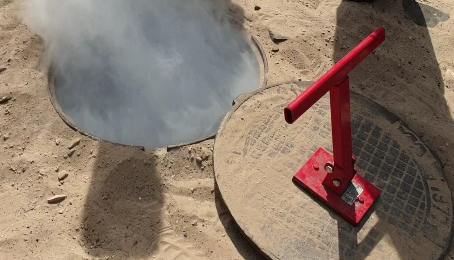 Atvērta kanalizācijas akas lūka smilšainā teritorijā; no akas paceļas balti dūmi vai tvaiks, bet blakus novietots noņemts vāks ar sarkanu rokturi.