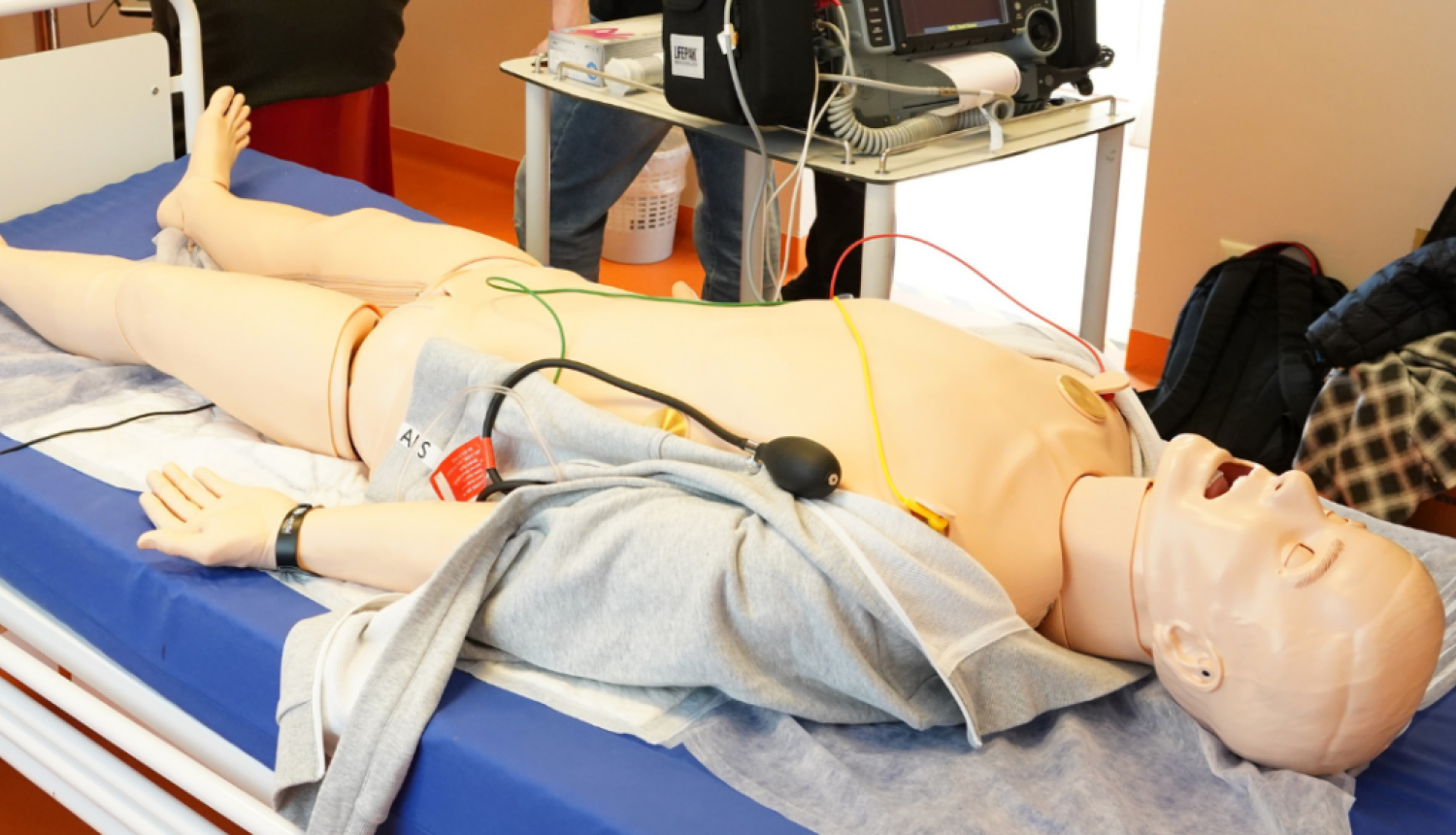 Mācību telpā uz gultas novietots medicīnas simulators ar pieslēgtiem vadiem un monitorēšanas aprīkojumu, kamēr vairāki cilvēki fonā vēro informāciju uz ekrāna.