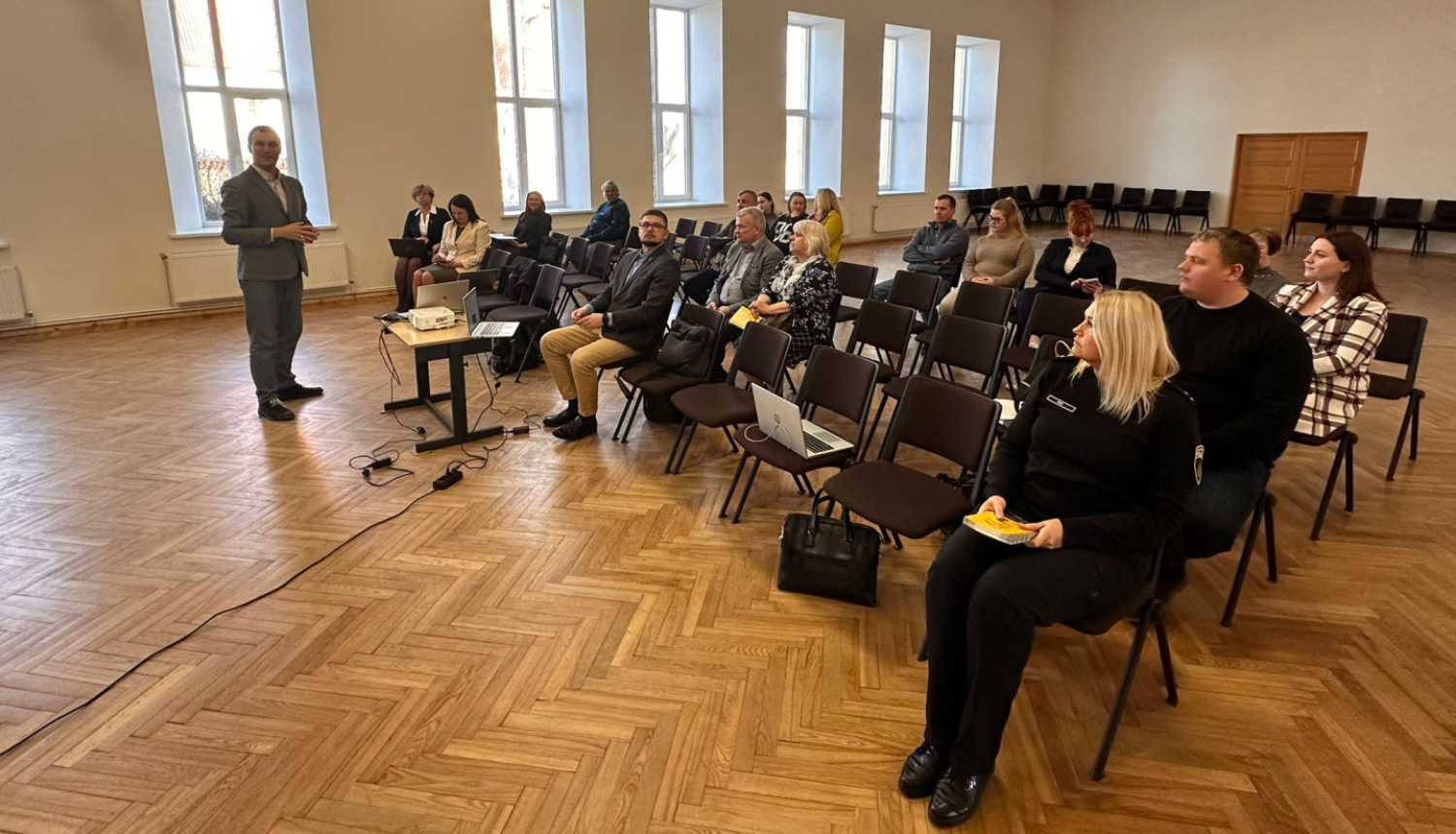 Laucienes kultūras nama zālē notiek Talsu novada pašvaldības tikšanās ar iedzīvotājiem. Priekšplānā redzams runātājs pie projektora galdiņa, bet zālē rindās sēž sanāksmes apmeklētāji un pašvaldības pārstāvji. Plašajā telpā ir gaišas sienas, augsti logi un koka grīda.