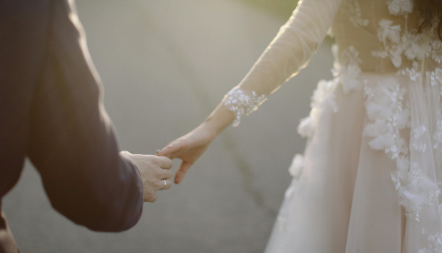 Tuvplānā redzamas divu cilvēku rokas, kas saņemas, vienam esot svinīgā uzvalkā, otram – baltā kāzu tērpā.