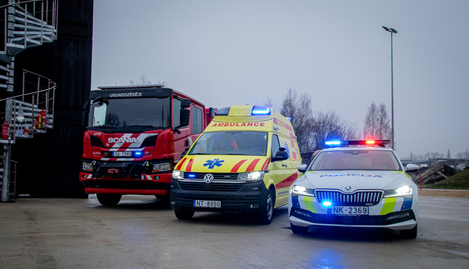 Attēlā redzami trīs operatīvie transportlīdzekļi – ugunsdzēsēju automašīna, Neatliekamās medicīniskās palīdzības dienesta automašīna un Valsts policijas dienesta auto – novietoti blakus uz laukuma. Visām automašīnām ieslēgtas bākugunis. Fonā redzama ēka, laterna un koki apmākušās debesīs.