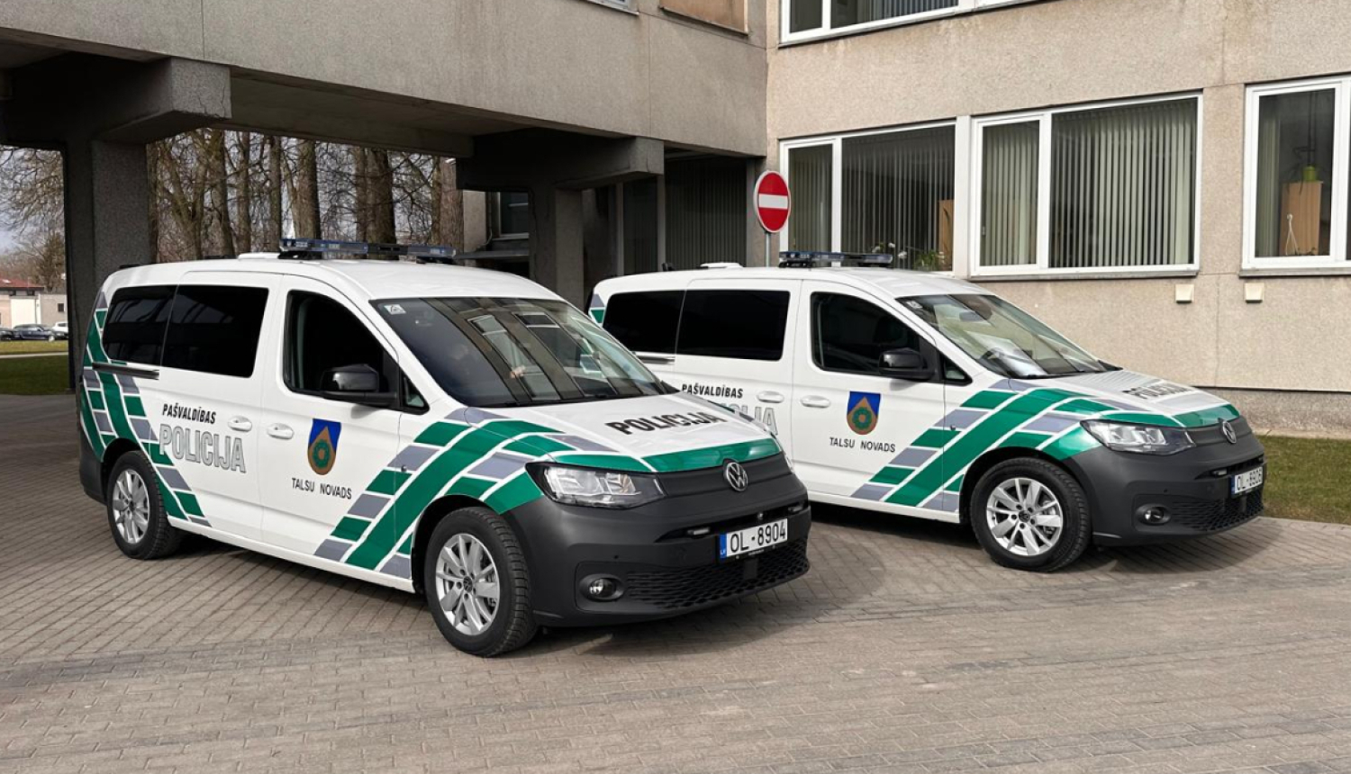 Attēlā pie administratīvās ēkas novietoti divi jauni Talsu novada Pašvaldības policijas operatīvie transportlīdzekļi – balti Volkswagen Caddy ar zaļi pelēku trafarējumu, uzrakstiem „Pašvaldības policija” un Talsu novada simboliku. Abiem transportlīdzekļiem uz jumta redzamas bākugunis.