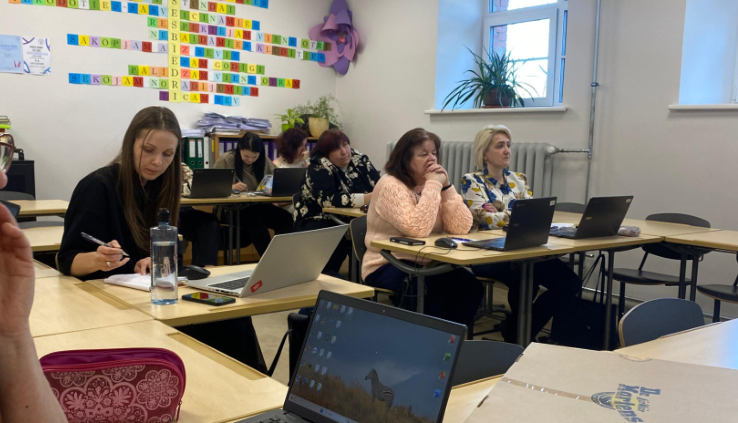 Klasē pie galdiem sēž pedagogi ar klēpjdatoriem un piezīmēm, piedaloties darbseminārā. Priekšplānā redzams galds ar datoru, krūzi un darba materiāliem, bet fonā – dalībnieki, kas klausās un strādā pie uzdevumiem.