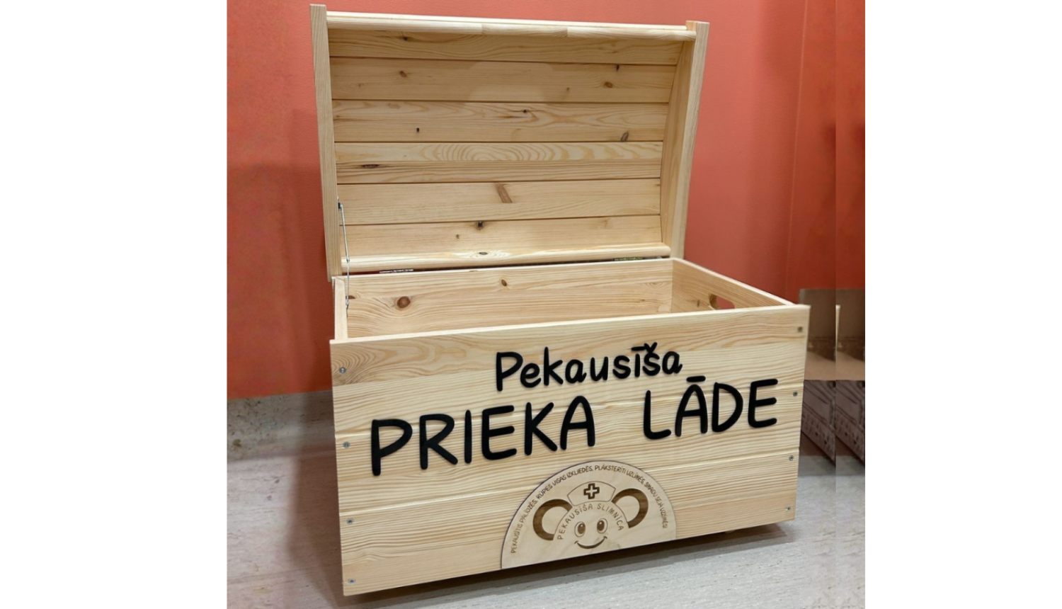 Atvērta koka kaste ar uzrakstu „Pekausīša prieka lāde”, novietota uz galda pie oranžas sienas. Uz kastes redzams arī Pekausīša slimnīcas logo.