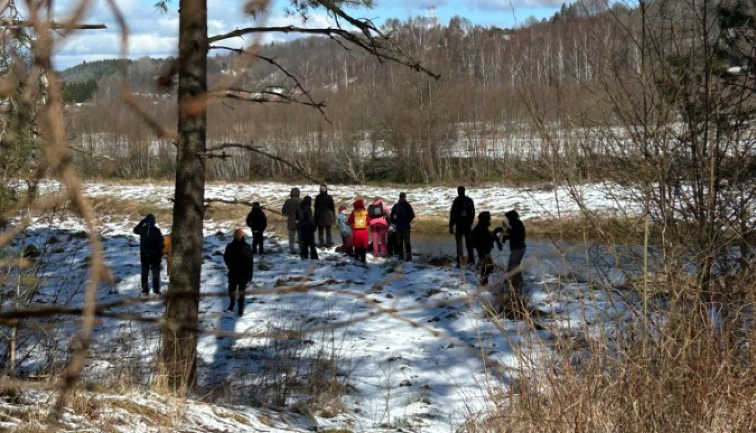 Cilvēku grupa agrā pavasara dienā stāv sniegainā upes vai dīķa krastā, fonā redzami kaili koki un zilas debesis ar mākoņiem. Skats fotografēts no neliela uzkalna caur koku zariem, priekšplānā redzama iemīta taka.