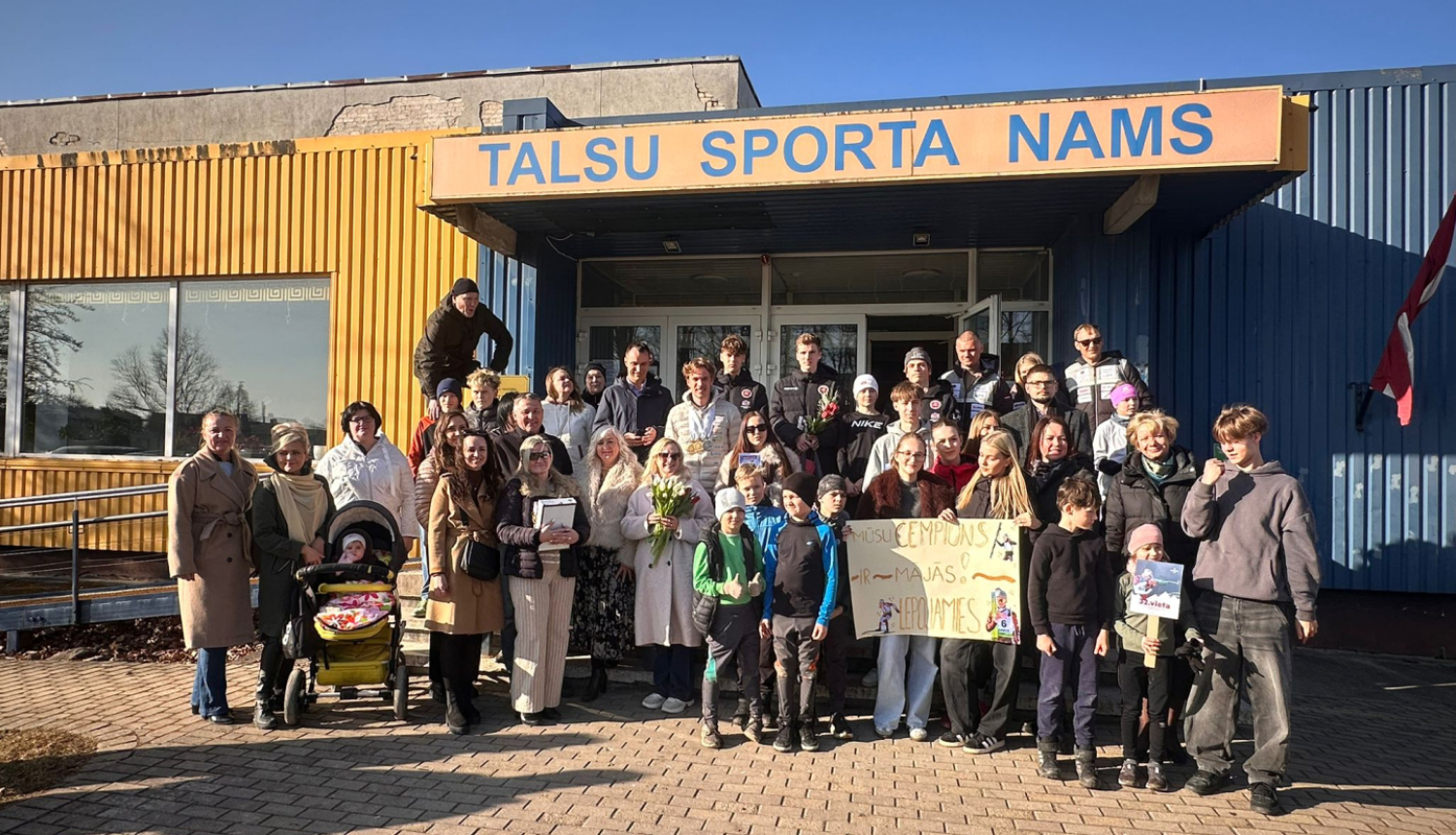 Pie ēkas ieejas redzama cilvēku grupa, kas svinīgi sagaida sportistu, turot rokās apsveikuma plakātu.