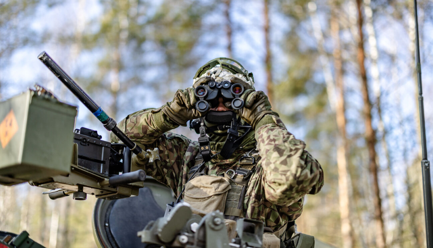 Mežainā apvidū redzams karavīrs maskēšanās formā un ekipējumā, kurš caur binokli vēro apkārtni. Viņš atrodas pie militārās tehnikas aprīkojuma, bet fonā redzami koki un skaidras debesis.