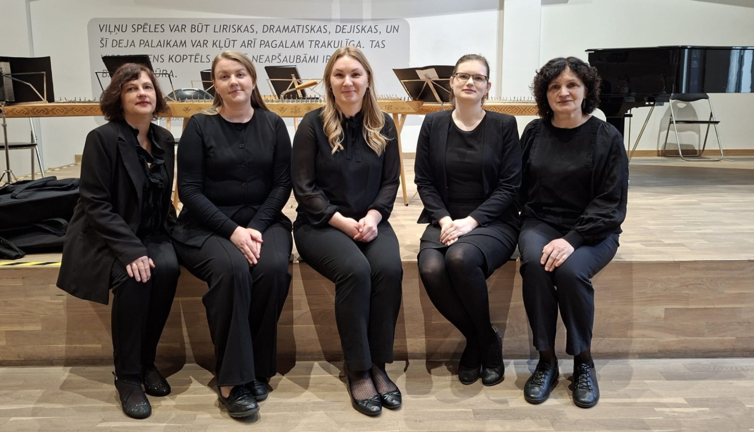Attēlā redzamas piecas sievietes melnā koncerttērpā, sēžot uz skatuves malas. Fonā redzamas kokles, nošu pultis un flīģelis, kas liecina par muzikālu uzstāšanos vai koncertu.