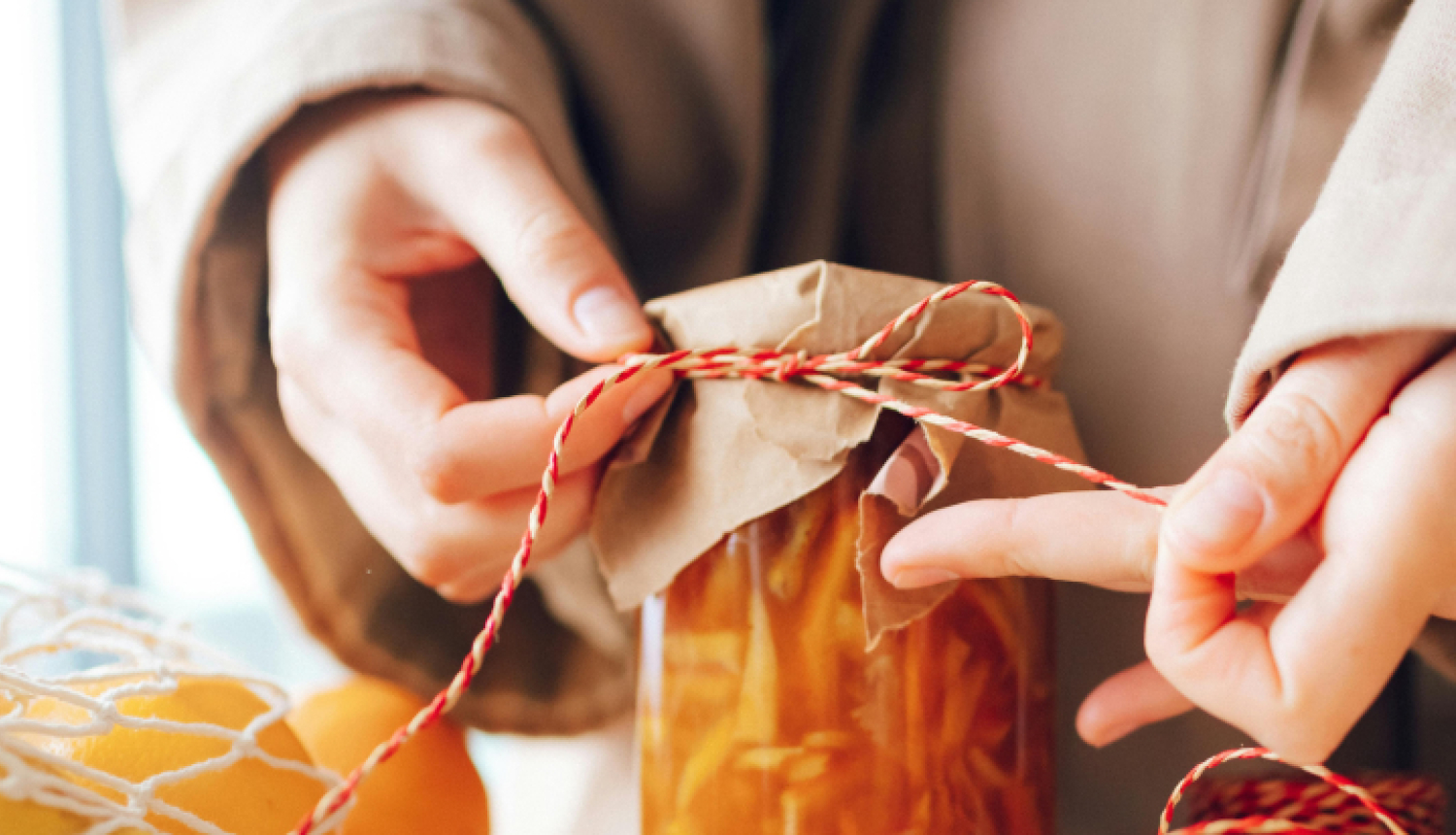 Tuvplānā redzamas rokas, kas apsien aukliņu ap stikla burciņu ar ievārījumu vai sīrupu. Burciņa pārklāta ar brūnu papīru, bet blakus uz galda atrodas citrusaugļi, koka karote un sarkani balta aukla, radot siltu, mājīgu noskaņu.