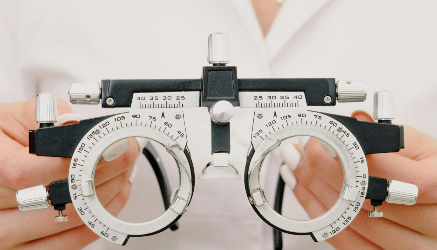 Optometrists tur rokās redzes pārbaudes ierīci (foropteru) ar regulēšanas skrūvēm un graduētām skalām, gatavojoties noteikt briļļu lēcu stiprumu.