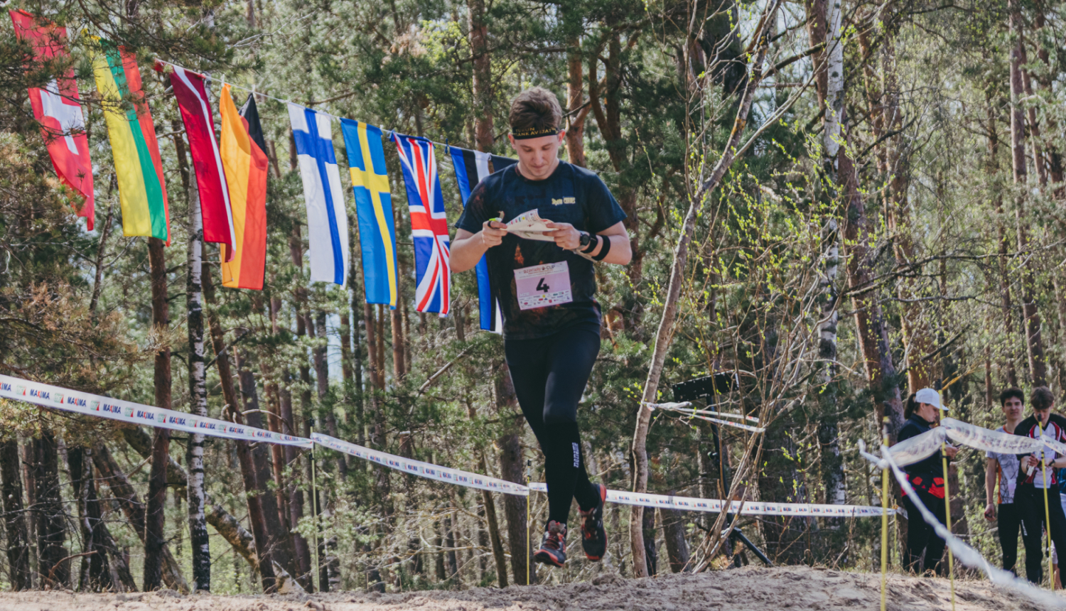 Orientierists skrien pa smilšainu meža posmu, lasot karti; fonā starp kokiem izkārti dažādu valstu karodziņi un redzami sacensību norobežojumi.