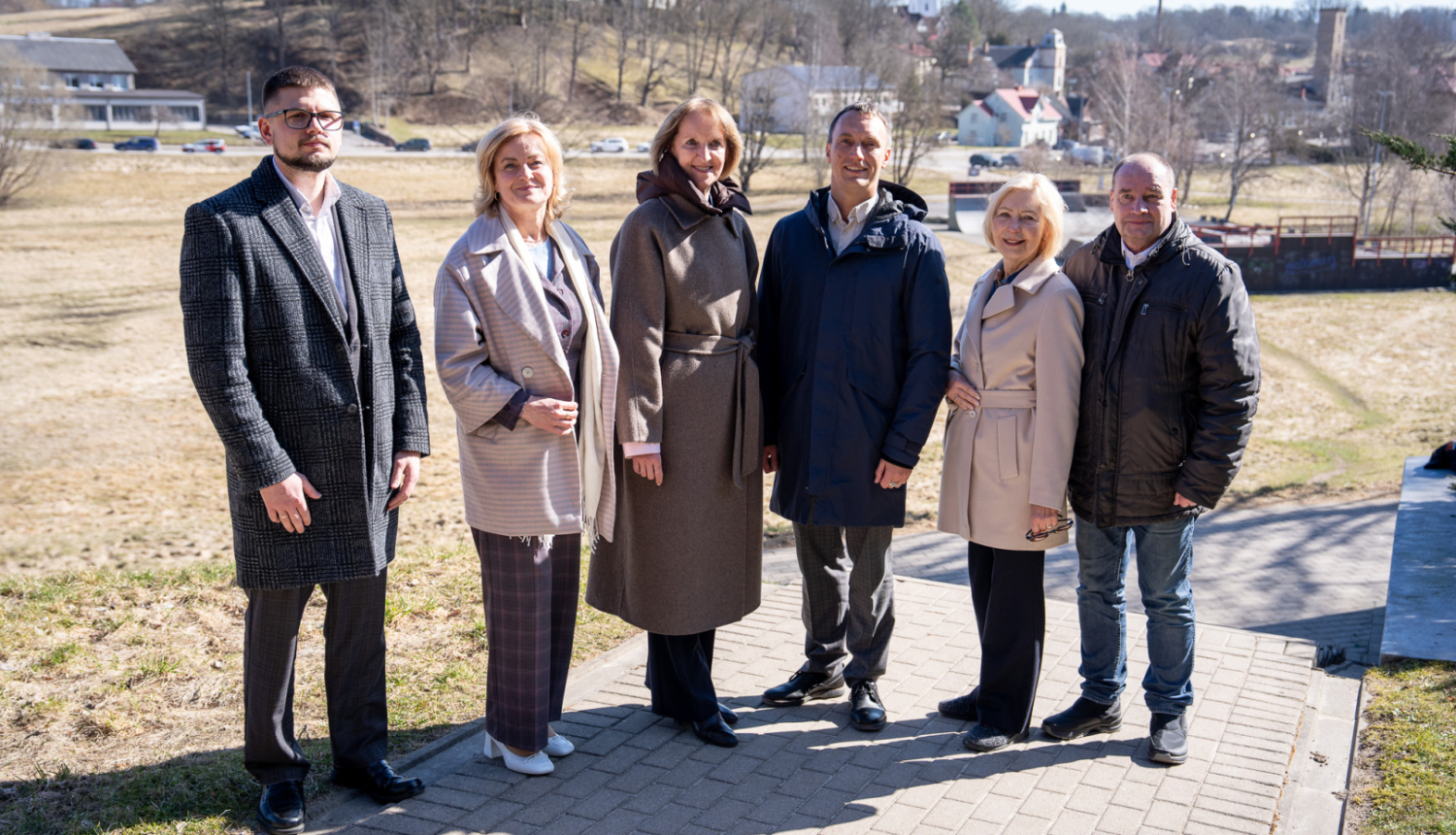 Alt teksts:  Pieci cilvēki un viens jaunietis stāv ārā uz bruģēta celiņa saulainā pavasara dienā. Fonā redzama Talsu ainava ar pakalniem, kokiem un pilsētas ēkām, tostarp baznīcas torņiem. Grupa pozē kopīgai fotogrāfijai, visi ģērbušies mēteļos un virsjakās.