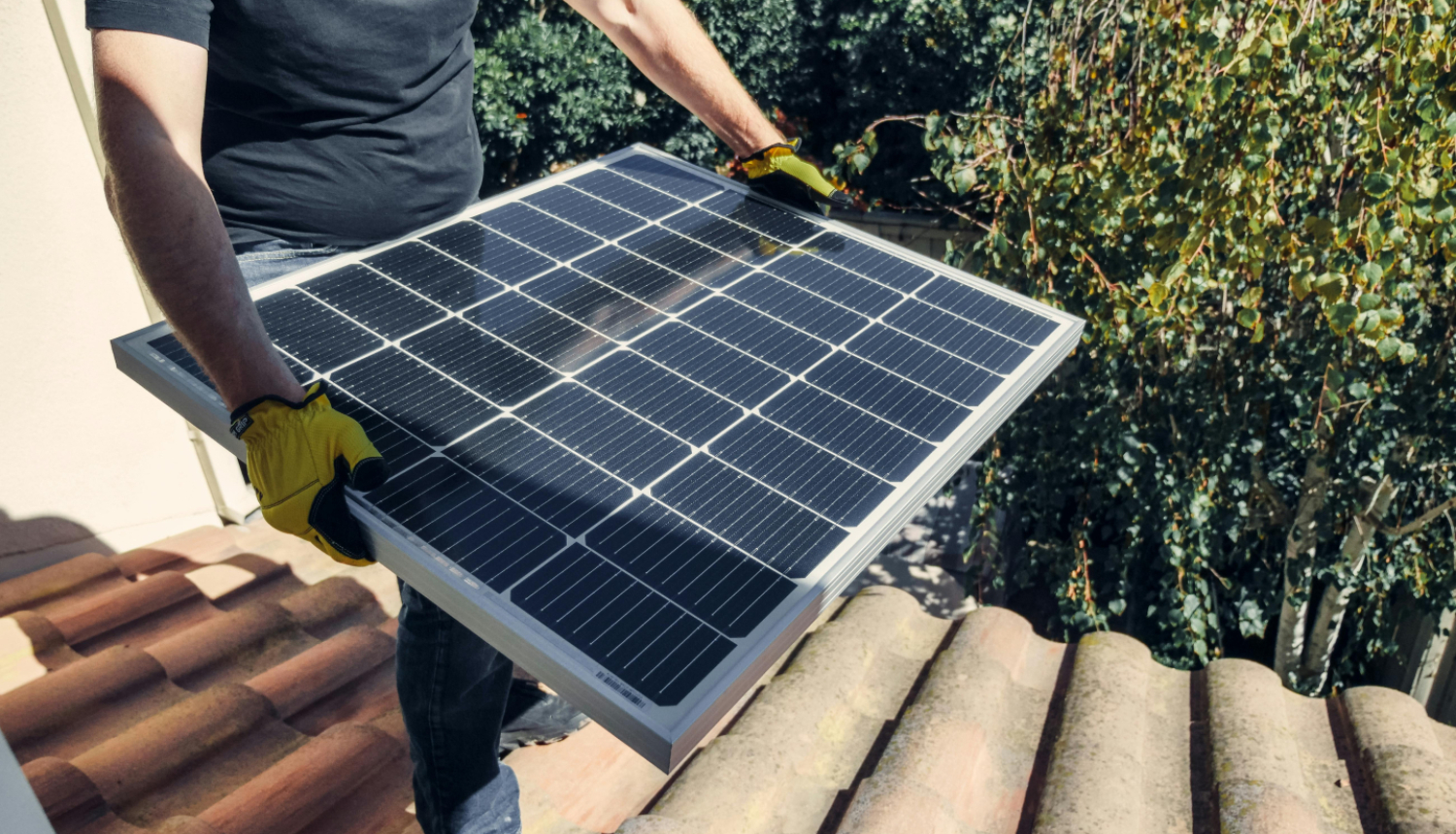 Attēlā redzams cilvēks, kurš uz ēkas jumta tur rokās saules paneli, gatavojoties tā uzstādīšanai. Fonā redzami koki un saulains laiks.