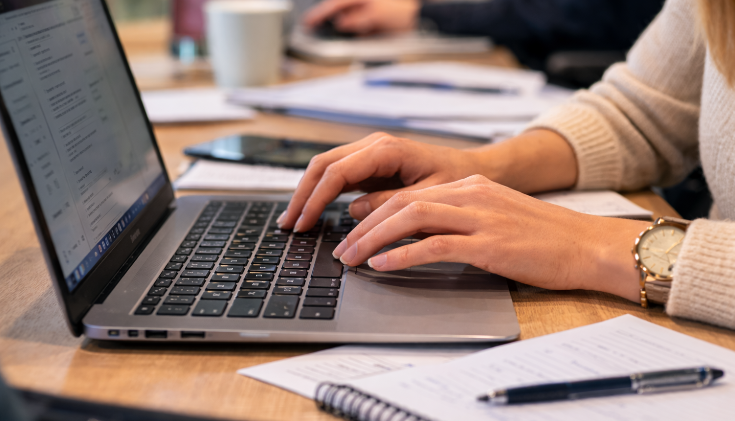 Tuvplāna foto redzamas rokas, kas raksta uz klēpjdatora tastatūras pie darba galda; blakus novietota piezīmju klade un pildspalva, fonā izplūdis biroja darba process.