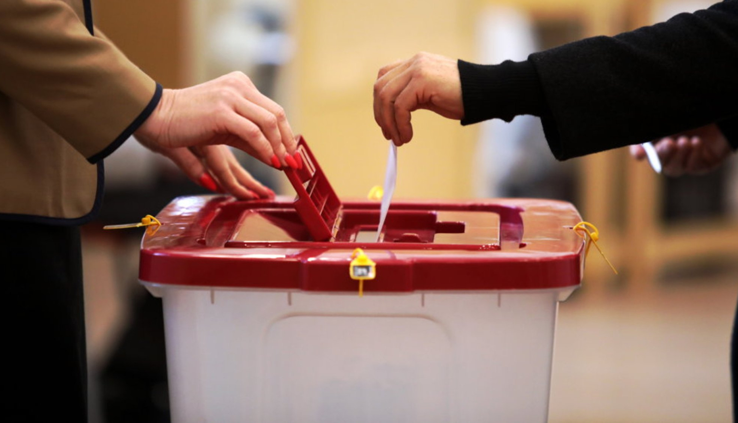Tuvplānā redzama balsošana vēlēšanu iecirknī – divi cilvēki vienlaikus ievieto vēlēšanu zīmes plastmasas balsošanas kastē ar sarkanu vāku. Uz kastes redzamas drošības plombas, bet fonā izplūdusi iekštelpu vide.